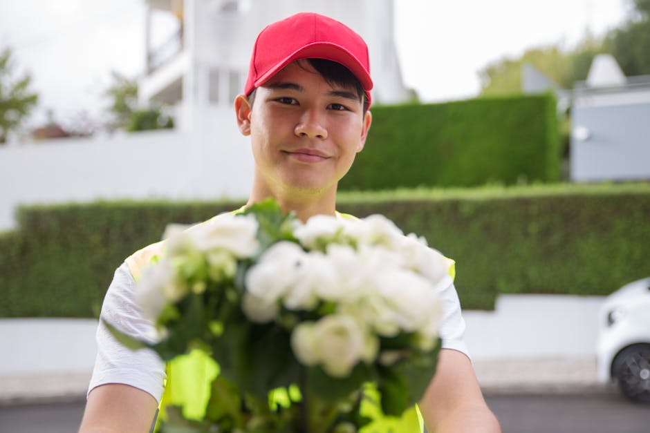 A man dressed in a red cap and red jacket holding a colorful bouquet of fresh flowers wrapped in patterned paper. The bouquet features vibrant yellow, blue, pink, and red flowers with lush green folia