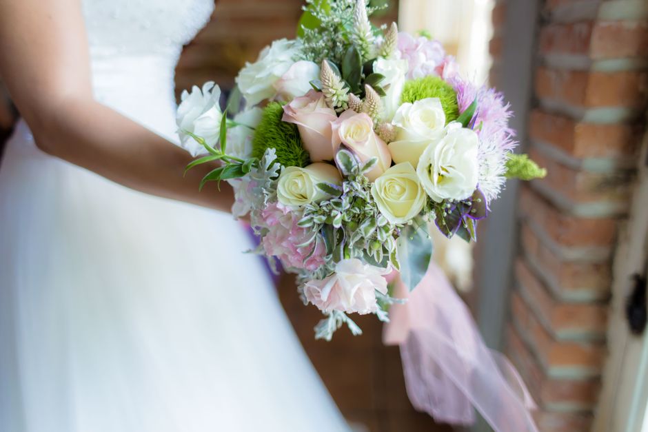 A bride is holding a floral bouquet arrangement consisting of creamy white roses, light pink hydrangeas, and soft green foliage, accented with purple-tinted leaves and small white flowers, wrapped wit