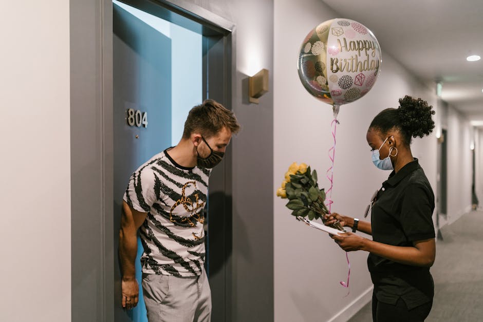 A woman wearing a face mask and black uniform is holding a bouquet of yellow roses, prepared for flower delivery, while standing in a hallway with grey walls and a grey carpet. She presents the floral