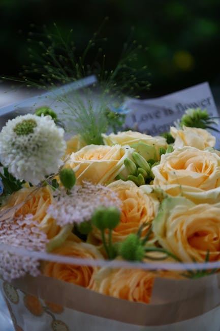 A floral arrangement featuring pale peach roses with tightly packed, velvety petals surrounded by small white pom-pom chrysanthemums, delicate green buds, and wispy green foliage. The bouquet is wrapp