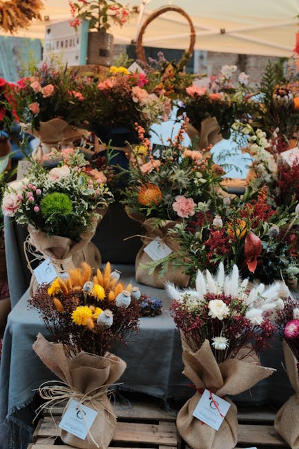 A display of various floral arrangements at a market stall, featuring bouquets wrapped in brown craft paper and tied with twine. The arrangements include a mix of flowers such as pink roses, white dai