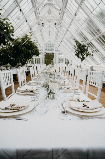 A long dining table set for a formal event inside a bright, glass-walled conservatory with a high, arched ceiling. The table is covered with a white tablecloth and decorated with a centerpiece of deli