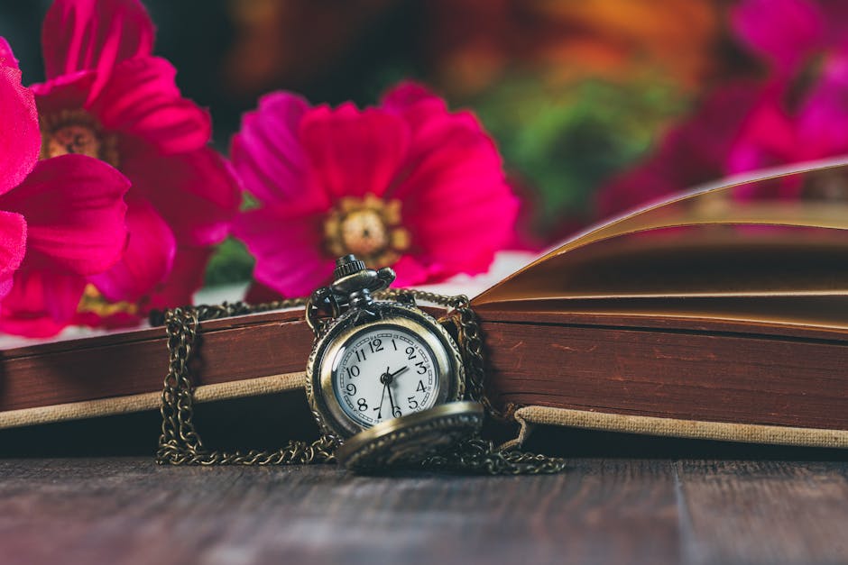 A close-up view of a vintage silver pocket watch with a white dial and black numerals, draped over an open hardcover book with yellowed pages. The book rests on a dark wooden surface, with bright pink